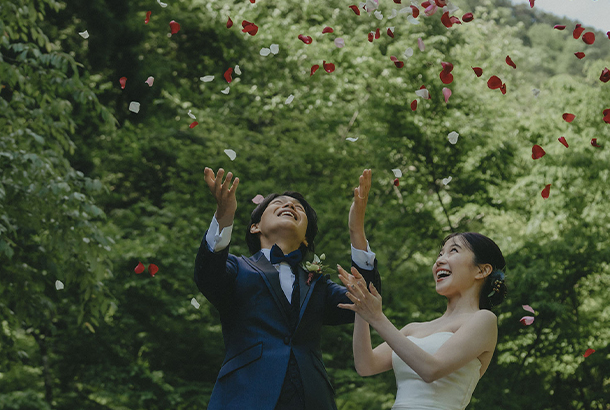 写真：洋装の婚礼衣装を身にまとった新郎新婦の周りにバラの花弁が舞っている